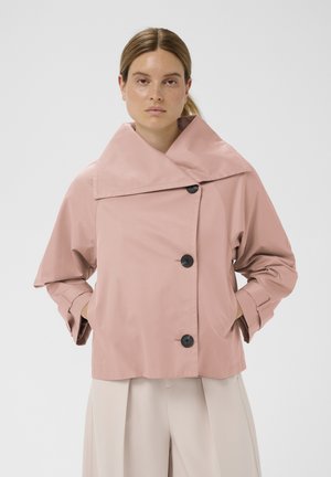 Femme portant une veste ample rose à grand col et boutons noirs, mains dans les poches, associée à un pantalon beige clair à jambes larges, sur fond blanc.
