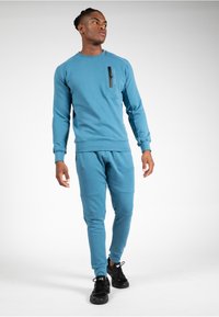 Turquoise sweatshirt met ritszak op de borst, gecombineerd met bijpassende tapered joggers. Beide zijn gemaakt van zachte, gestructureerde stof en hebben ribgebreide boorden.