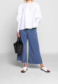 Camicia bianca con maniche a palloncino e bottoni sul davanti, abbinata a jeans blu a gamba larga. Borsa nera con accenti di perle e sandali bianchi.