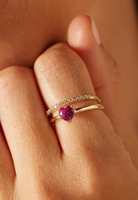 Twee ringen aan een hand: de ene met een roze hartvormige edelsteen, de andere met kleine heldere stenen, beide gezet in een gouden band.