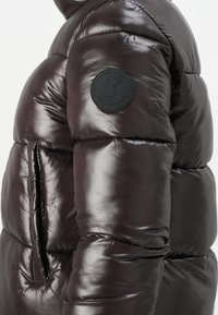 Close-up van een glanzende donkerbruine gewatteerde jacksleeve met een ronde "Save The Duck" logo patch.