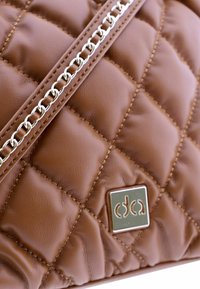 Bolso de mano de cuero marrón acolchado con un detalle de cadena plateada y una placa de logo metálica. Textura suave y diseño acolchonado con costuras visibles.