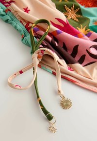 Tissu coloré de maillot de bain avec motif d'étoiles de mer, bord élastique froncé et bretelles nouées terminées par de petites breloques soleil dorées sur une surface blanche.