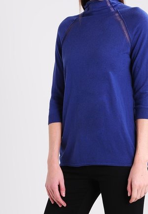 Kvinde iført en langærmet, kongeblå rullekravesweater med sorte bukser, stående mod en ensfarvet lys baggrund.
