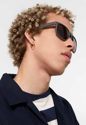 Giovane con capelli ricci che indossa occhiali da sole neri, orecchino a cerchio dorato, giacca blu navy e camicia a righe, guarda di lato.