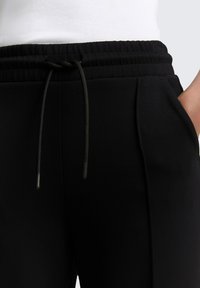Schwarze Jogginghosen mit elastischem Bund, verstellbarem Kordelzug, Seitentaschen und glatter Textur. Eine Nahaufnahme hebt das Design des Bundes hervor.