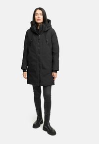 Sort parka med lynlås foran, hætte og justerbare snore. Lommer foran med en glat, mat tekstur. Båret over sorte jeans og støvler.