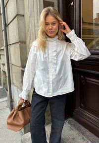 White cotton blouse with ruffled lace collar and cuffs, paired with dark jeans. Holding a brown leather handbag. External stone wall background.