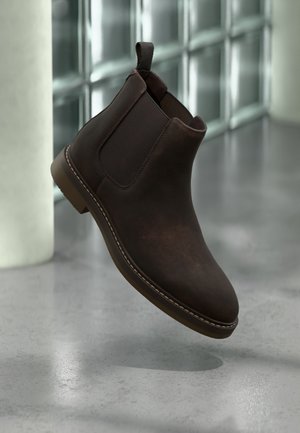 Bottine Chelsea en cuir marron avec des panneaux latéraux élastiques et un onglet de traction, flottant au-dessus d'un sol en béton poli près d'un mur en blocs de verre.