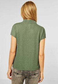 Grön kortärmad polo med krage, mjuk textur och avslappnad passform. Bakvyn visar släta sömmar och diskreta detaljer på plagget.