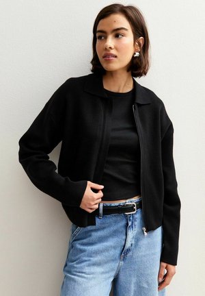 Cardigan noir zippé avec un design à col. Matériau léger, coupe ample, associé à un haut noir et un jean bleu taille haute.