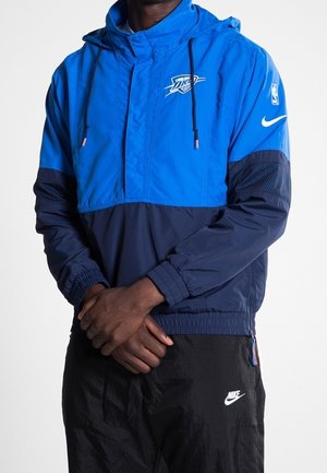 Personne portant une veste coupe-vent Oklahoma City Thunder Nike bleu et marine et un pantalon Nike noir, debout les mains jointes.