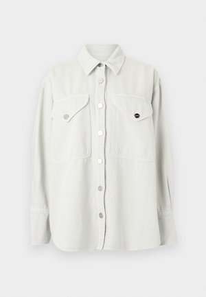 Femme portant une chemise blanche oversize, un jean beige à jambes larges, un top blanc, des bottes marron, une ceinture jaune à boucle, tenant un grand sac marron foncé.