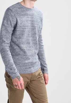 Uomo che indossa un maglione grigio a maglia e pantaloni marroni, mostrando la mano sinistra tatuata con una farfalla e simboli sulle dita.