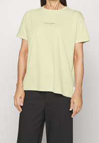 Světlé zelené bavlněné tričko s kulatým výstřihem a krátkými rukávy. Na střední přední straně je minimální nápis. Volný střih, v kombinaci s černými kalhotami.