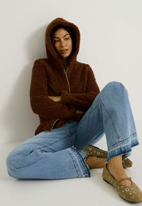 Haut à capuche zippé marron en peluche, associé à un jean large déchiré bleu clair. Les chaussures sont beiges avec des découpes circulaires.