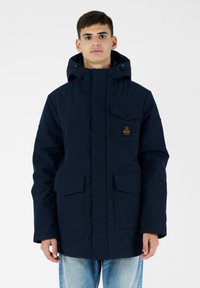 Parka blu navy con cappuccio staccabile, chiusura frontale con zip, multiple tasche con flap e patch con logo sul petto. Tessuto dalla texture liscia.