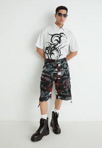 Weißes kurzärmeliges Hemd mit schwarzem Grafikdesign, kombiniert mit camo Cargo-Shorts in Grau, Rot und Schwarz, und schwarzen Kampfstiefeln mit weißen Socken.