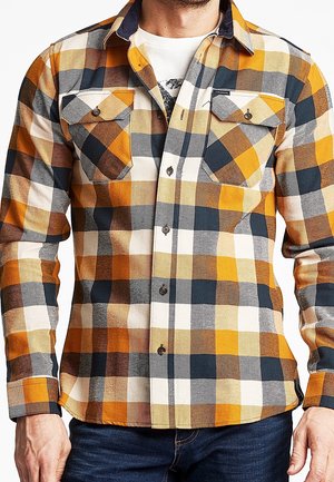Homme portant une chemise à manches longues à carreaux jaune, marron, noir et blanc par-dessus un T-shirt blanc avec un jean bleu foncé.