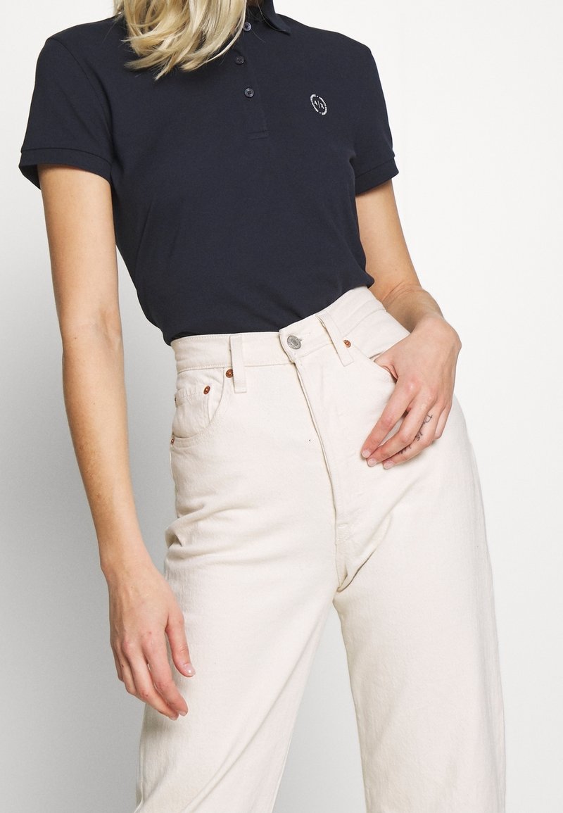 En kvinna poserar iklädd en marinblå polo med en logotyp och krämfärgade högmidjade jeans, med handen på höften, mot en ljus bakgrund.