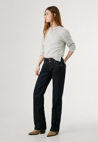 Svetlo siva pletena pulover s dolgimi rokavi, v kombinaciji s temno modrimi kavbojkami ravnega kroja, ki imajo gumbe in vidno šivanje.