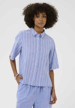 Žena s kučeravými vlasmi, oblečená v modro-bielej vertikálne pruhovanej krátkemu rukávu so zapínaním na gombíky a rovnako modrých nohaviciach, stojaca s rukou v vrecku.