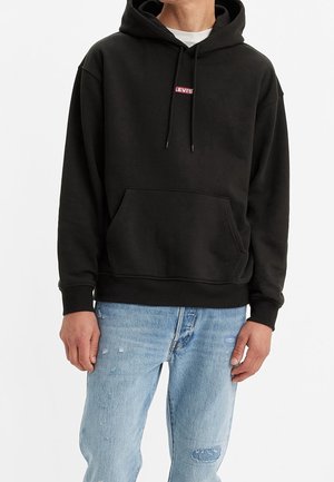 Person iført en sort Levi's hættetrøje med frontlomme og snore, kombineret med lyseblå slidte jeans.