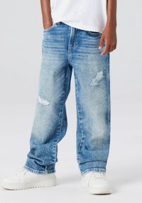 Jeans in denim azzurro chiaro leggermente consumati con vestibilità ampia, dotati di tasche frontali e dettagli sfrangiati sulle ginocchia, abbinati a sneakers bianche.