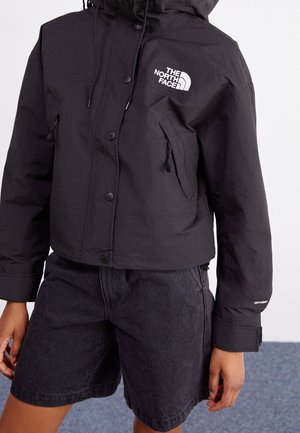 Personne portant une veste noire North Face avec des boutons sur le devant et des poches latérales zippées, assortie à un short en jean noir.