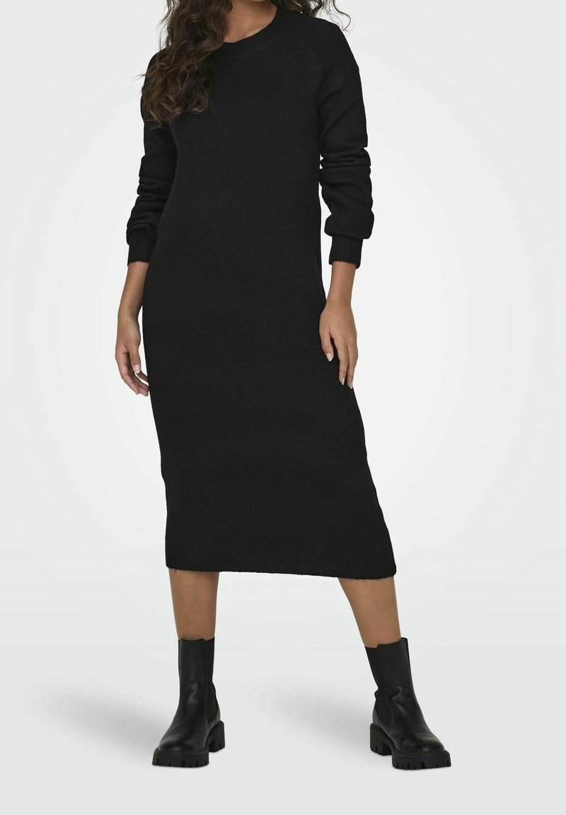 Femme portant une longue robe noire en maille à manches longues et des bottines noires, debout devant un fond clair uni.