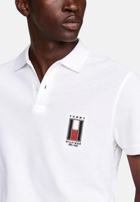 Camisa polo blanca con un acabado texturizado, que presenta un pequeño logo bordado de Tommy Hilfiger en azul, rojo y blanco en el pecho.
