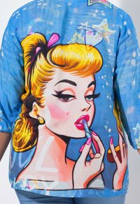 Femme aux cheveux blonds et aux ongles roses appliquant du rouge à lèvres, sur fond bleu avec des taches blanches et des détails d'étoiles de mer.