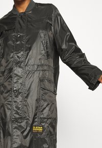 Svart glänsande lång jacka med knäppning, flera fickor och en gul G-Star Originals Raw Denim-logotyp på nedre fronten.