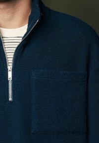 Dunkelblauer Fleece-Pullover mit einem Viertel-Reißverschluss-Kragen, ausgestattet mit einem strukturierten Stoff und einer aufgesetzten Tasche vorne, über einem gestreiften Hemd getragen.