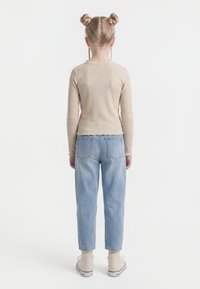 Bērns ar blondiem matiem divās bungās, valkājot bēša krāsas garām piedurknēm džemperi, gaiši zilas džinsas un baltas sporta kurpes, stāvot ar muguru pret baltu fonu.