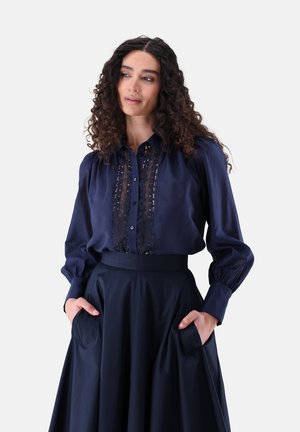 Frau mit lockigem Haar, die eine marineblaue Bluse mit Spitzendetails und einen hoch taillierten marineblauen Rock trägt, Hände in den Taschen, schaut leicht nach links.