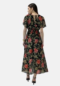 Vestido maxi de tela negra transparente, con un estampado floral en rojo y verde. Cintura elástica y espalda descubierta con detalle de lazo. Tacones negros.
