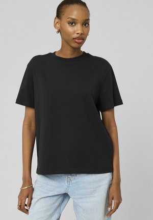 Femme portant un t-shirt noir uni à manches courtes, un jean bleu clair, des boucles d’oreilles anneaux dorées et des bracelets, debout devant un fond gris.