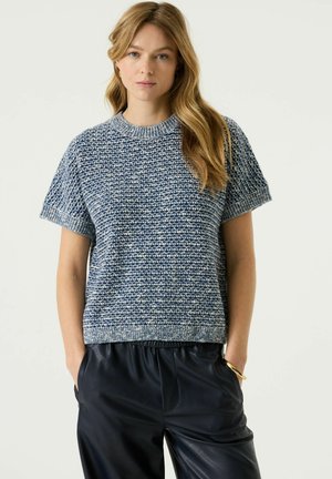 Vrouw met lang blond haar draagt een blauw-wit gebreide trui met korte mouwen en zwarte leren broek, staand met handen in de zakken.
