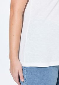 Top blanco de manga corta visto de lado, con una textura suave. Combinado con unos vaqueros de mezclilla azul claro, que muestran un ajuste relajado.