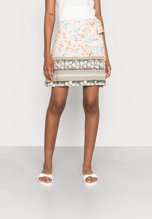 Mannequin portant une jupe portefeuille fleurie longueur genou avec des motifs pêche, bleu et vert, associée à des sandales blanches à enfiler et un haut blanc sans manches.