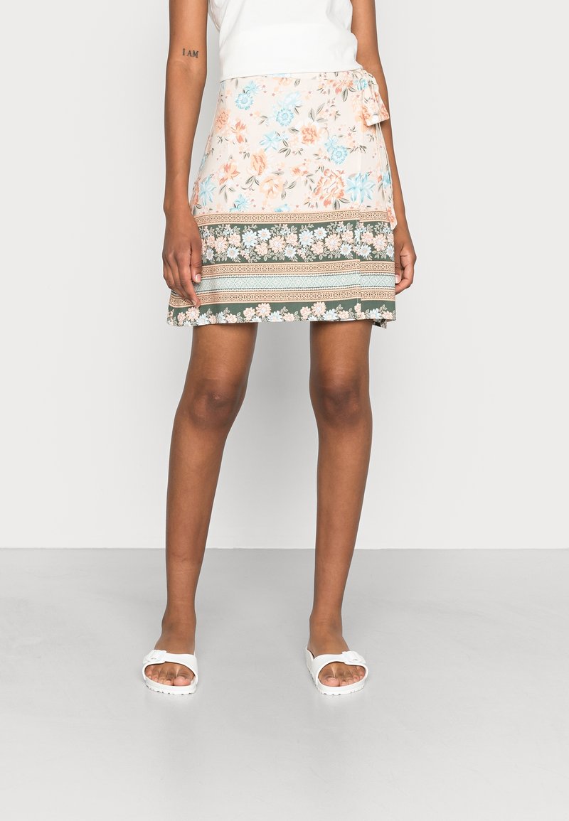 Mannequin portant une jupe portefeuille fleurie longueur genou avec des motifs pêche, bleu et vert, associée à des sandales blanches à enfiler et un haut blanc sans manches.