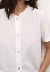 Camicia bianca in lino con maniche corte e scollo arricciato. Chiusura con bottoni di colore chiaro lungo la parte anteriore. Tessuto morbido.