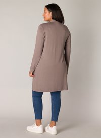 Vrouw draagt een lange taupekleurige cardigan, blauwe cropped spijkerbroek en witte platform sneakers, staand tegen een effen beige achtergrond.