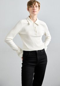 White ribbed long-sleeve polo shirt with a collar and button details, paired with high-waisted black jeans. Simple, structured design.