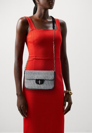 Kvinde iført en rød ærmeløs kjole med en sort-hvid mønstret crossbody-taske og et kraftigt guldarmbånd på håndleddet.