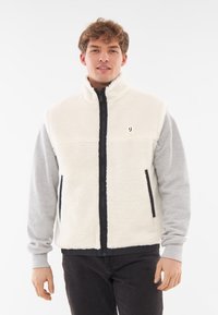 Fleeceväst i grädde med hög krage, full zip och grå ärmar. Har sidofickor och en liten logotyp på bröstet.