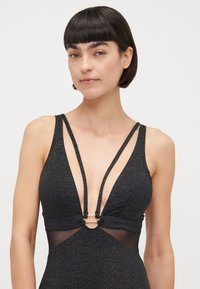 Μαύρο μαγιό με βαθύ V-ντεκολτέ, λαμπερή υφή και διάφα�νες πλαϊνές πάνελ. Διαθέτει λεπτές τιράντες και κυκλικό διακοσμητικό στη μέση.