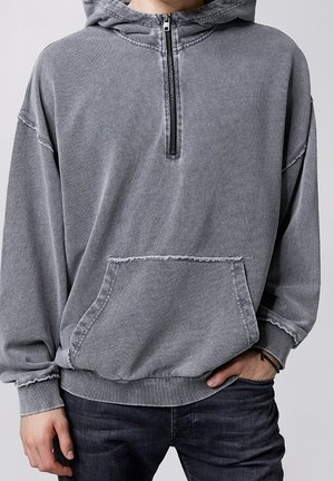 Persona che indossa una felpa con cappuccio grigia sbiadita con tasca frontale e chiusura con mezza zip, abbinata a jeans scuri.