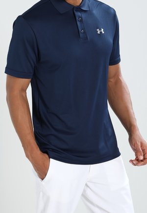 Homme portant un polo bleu foncé à manches courtes avec le logo Under Armour et un pantalon blanc, mains détendues le long du corps.